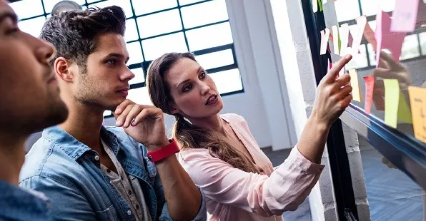 Zwei Personen betrachten ein Board mit Haftnotizen, eine Person zeigt mit dem Finger auf einzelne Inhalte.