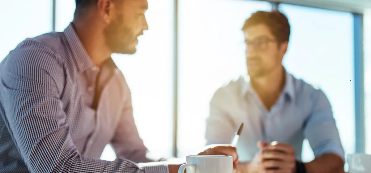Zwei Männer sitzen an einem Tisch, sprechen miteinander und haben eine Kaffeetasse und Schreibmaterial vor sich.