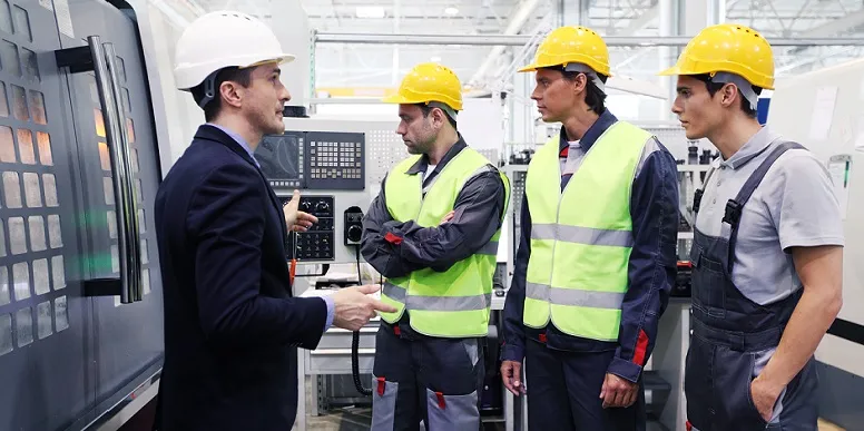 Vier Personen mit Schutzhelmen und Warnwesten stehen in einer Produktionshalle und führen ein Gespräch vor einer Maschine.