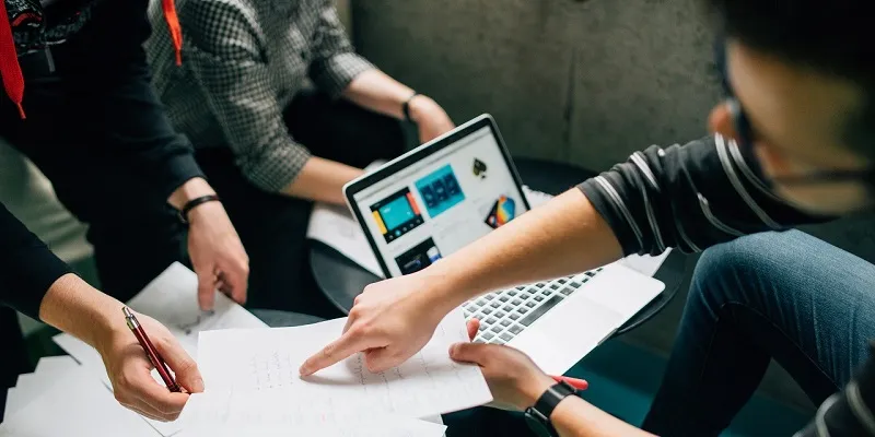 Mehrere Personen stehen um einen Tisch, zeigen auf ausgebreitete Unterlagen, im Hintergrund ist ein geöffnetes Notebook zu sehen.