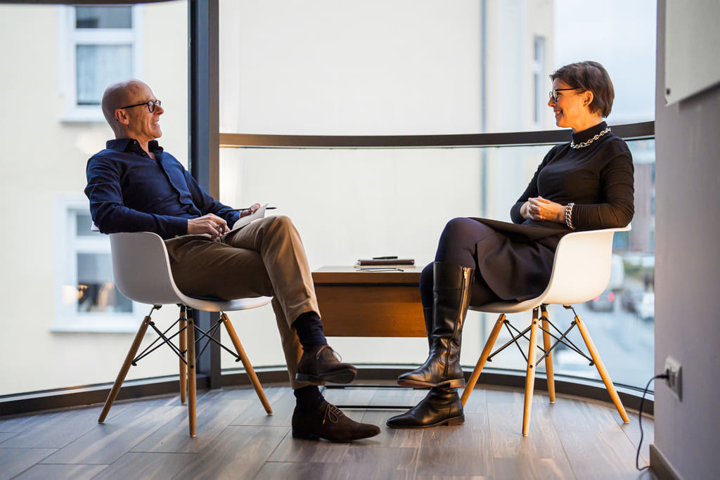 Zwei Personen sitzen sich in modernen Stühlen gegenüber und führen ein Gespräch in einem hellen Raum mit großer Fensterfront.