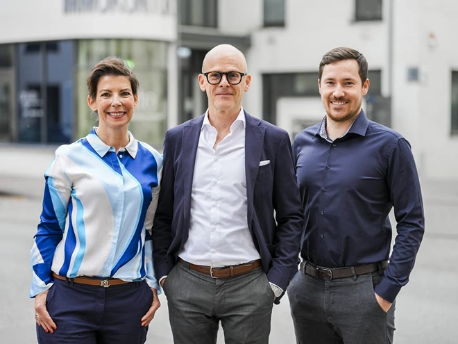 Rita Feilen, Ralph Schlüter und Jannik Rossmann stehen nebeneinander im Freien vor einem Gebäude. Sie sind in Smart-Casual-Kleidung gekleidet und lächeln in die Kamera.