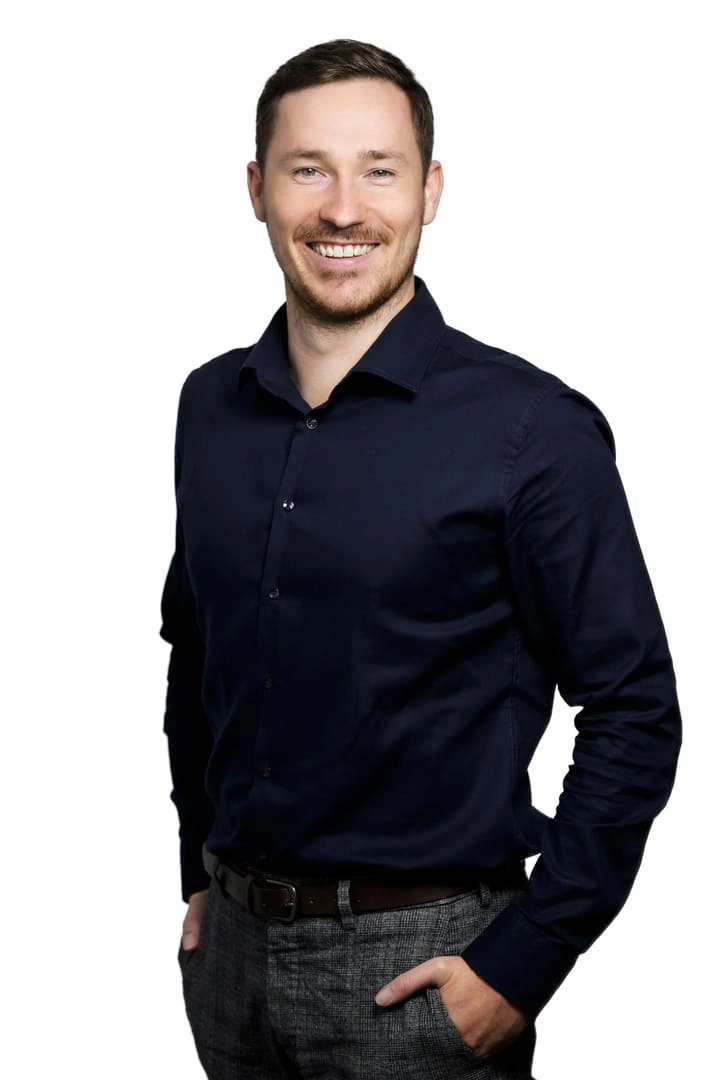 Porträt von Jannik Rossmann vor weißem Hintergrund. Er trägt ein dunkelblaues Hemd, eine graue Hose und lächelt freundlich in die Kamera.