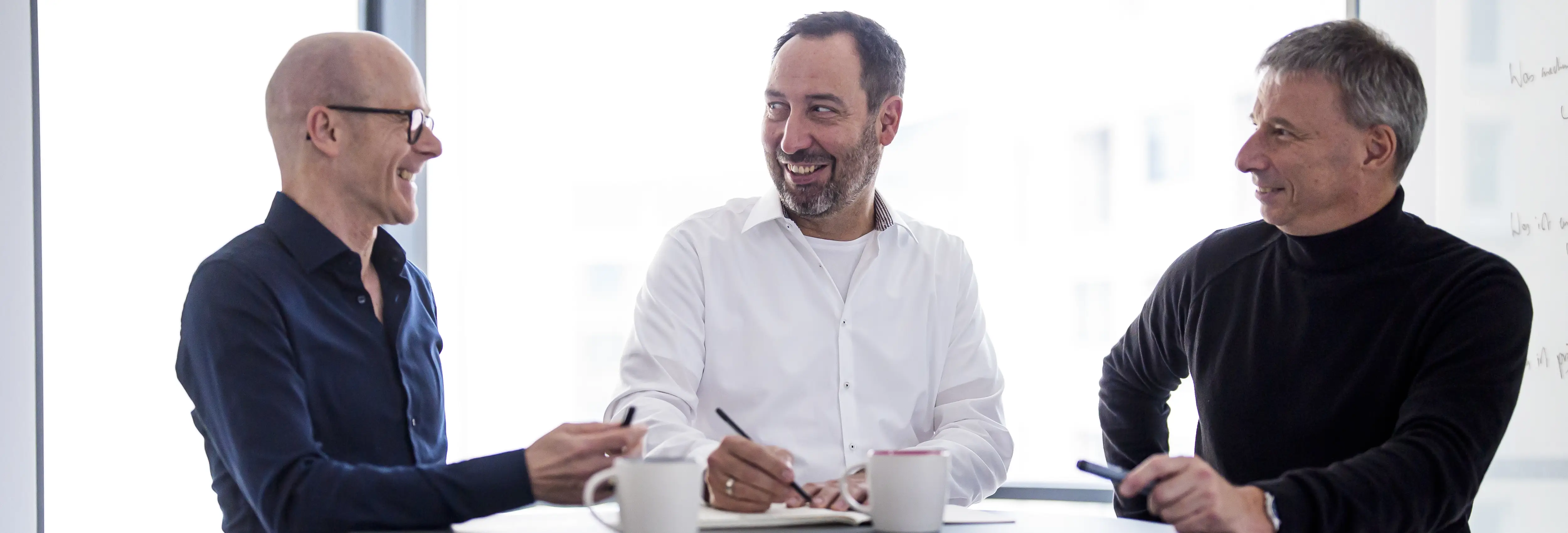 Drei Männer – Ralph Schlüter, Christoph Edlinger und Stefan Pollpeter – sitzen lächelnd an einem Tisch in einem hellen Büro. Sie führen ein entspanntes Gespräch, vor ihnen liegen Notizen und zwei Kaffeetassen.