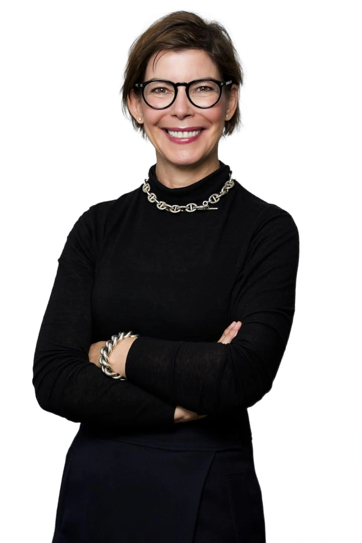 Porträt von Rita Feilen vor weißem Hintergrund. Sie trägt eine Brille, einen schwarzen Rollkragenpullover und eine auffällige Gliederkette. Sie hat die Arme verschränkt und lächelt.